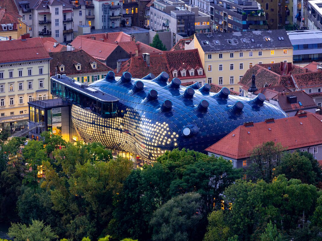 Futuristic dark blue building with round rooftop openings, surrounded by red tiled roofs and trees; modern architectural highlight in the middle of the historic old town. | © Graz Tourismus - Harry Schiffer