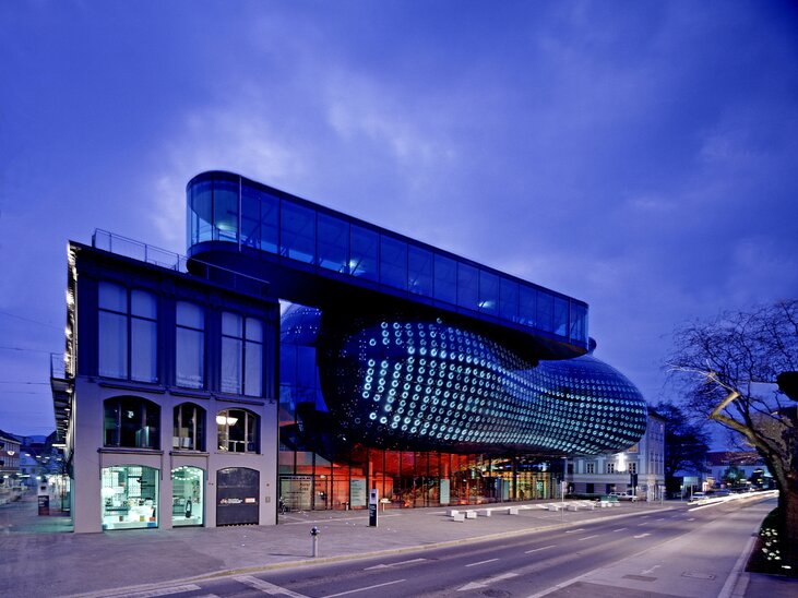 	Futuristic dark blue building with round rooftop openings, surrounded by red tiled roofs and trees; modern architectural highlight in the middle of the historic old town. | © Universalmuseum Joanneum