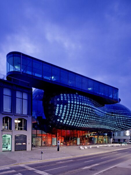 	Futuristic dark blue building with round rooftop openings, surrounded by red tiled roofs and trees; modern architectural highlight in the middle of the historic old town. | © Universalmuseum Joanneum