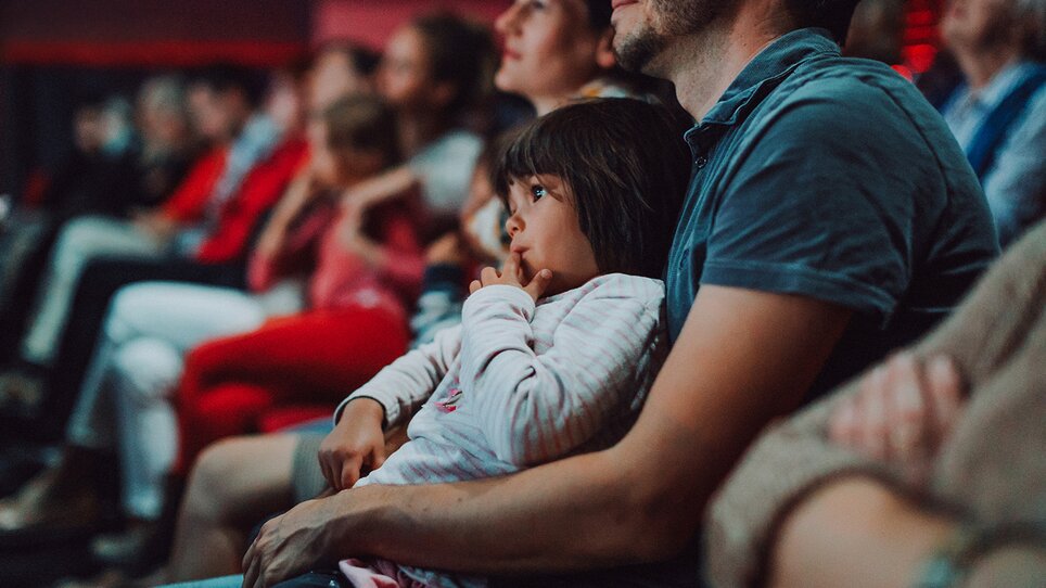 A group of people sits relaxed in Next Liberty. Children and adults watch the stage intently, surrounded by a friendly atmosphere. | © Stella