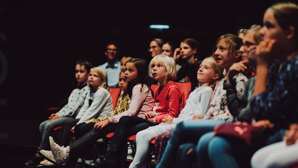 Children sit captivated in the Next Liberty venue. The dark room creates a focused atmosphere with flexible seating. | © Stella