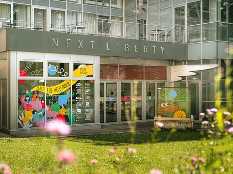 Das Next Liberty präsentiert moderne Architektur mit großen Fenstern und bunten Grafiken. Der Veranstaltungsort bietet eine helle Atmosphäre für Konferenzen und Networking. | © Stella