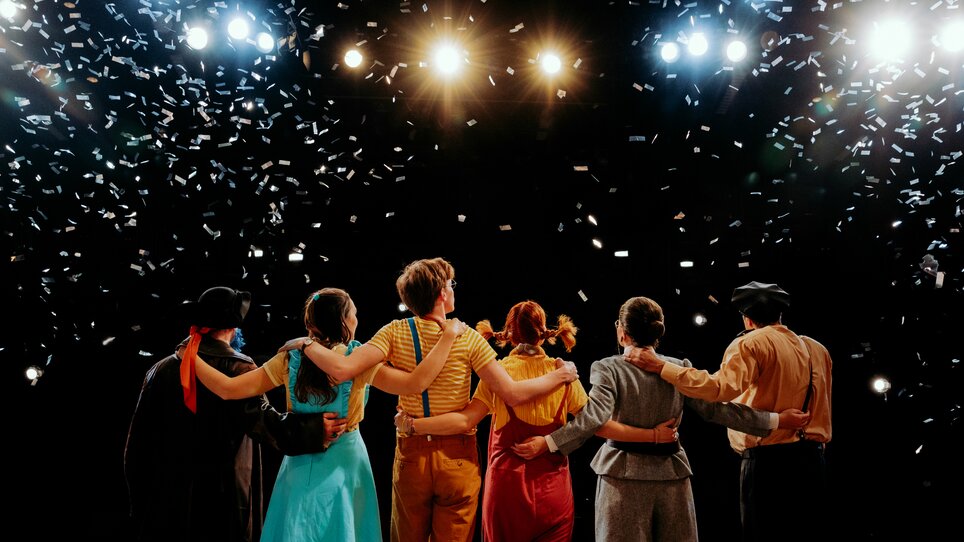The stage of Next Liberty features a group of performers during the final applause. Confetti falls and the stage lights create a celebratory atmosphere. | © Stella Kager