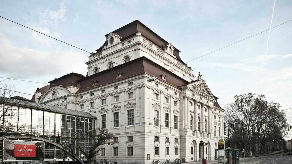 Oper Graz mit historischer Fassade, Glasbrücke und Haltestelle an leerer Straße | © Moodley Brand Identity