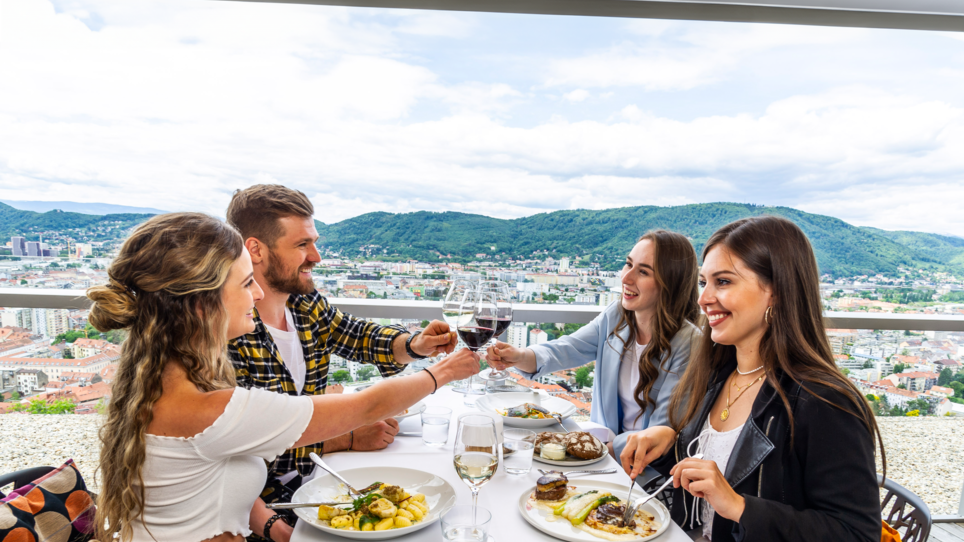 In Schlossberg Restaurant, four people sit around a table overlooking Graz. They toast with wine glasses and enjoy a stylish meal in a modern setting. | © Paul Stajan