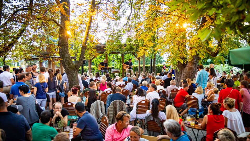 The Restaurant SCHLOSSBERG features a lively atmosphere with many guests. Guitarists perform on an outdoor terrace under trees. | © Restaurant SCHLOSSBERG - Werner Krug