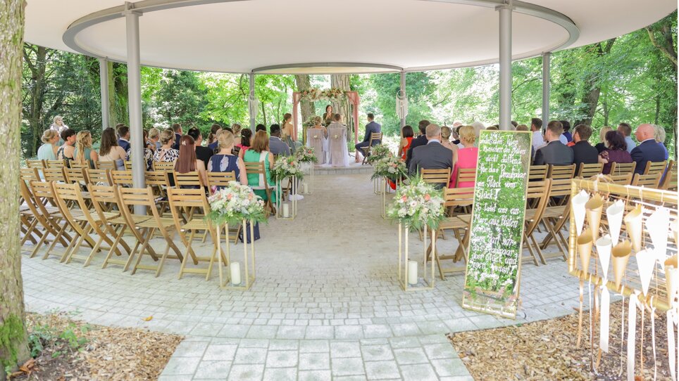 Couple at a ceremony surrounded by guests in a green setting. | © Sarah Raiser Fotografie
