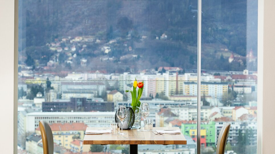 In Schlossberg Restaurant in Graz, a stylish table and two chairs offer a view of the city. Large windows provide an inspiring outlook on the Mur and surroundings. Modern design and ample lighting create a welcoming atmosphere for business trips and events. | © Paul Stajan