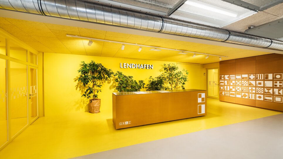 Der Lendhafen bietet einen hellen, modernen Empfangsbereich mit gelber Wandfarbe, einer großen Theke und Pflanzen. Der Raum fördert eine positive Atmosphäre für Veranstaltungen und Meetings. | © Lupi Spuma