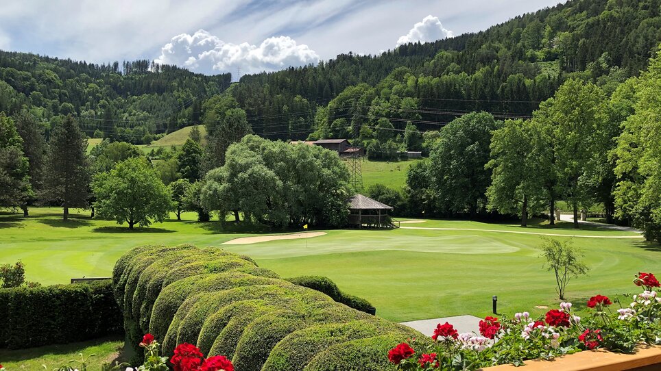 Green golf course surrounded by trees and flowers, mountains and clouds in the background next to Murhof Hotel & Restaurant. | © Murhof