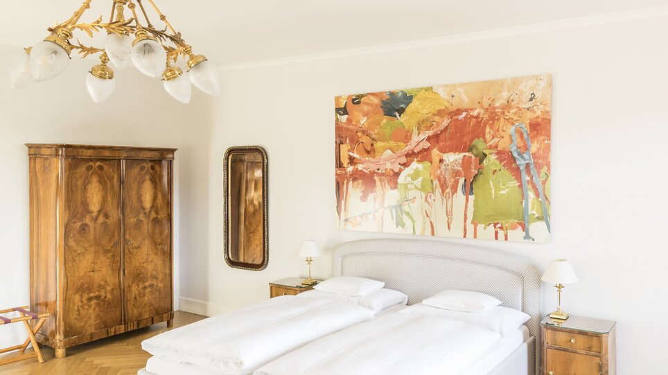 Bright bedroom with wooden wardrobe and abstract painting in Luminosa camera con due letti, opere d'arte e specchio nello Schlossberghotel. | © Schlossberghotel