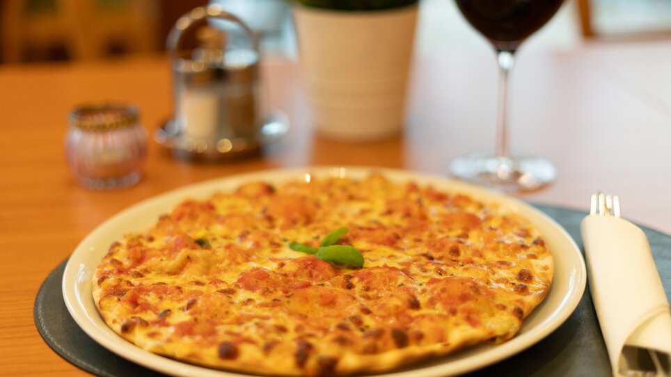 A plate of pizza with cheese and tomatoes, accompanied by a glass of red wine. | © Hotel Stoiser Graz