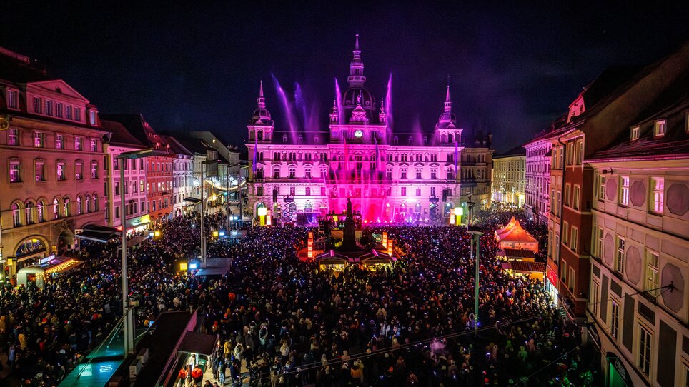 Blick auf die erleuchtete Stadt Graz mit vielen Menschen und einer Wasserfontäne. | © Erwin Scheriau