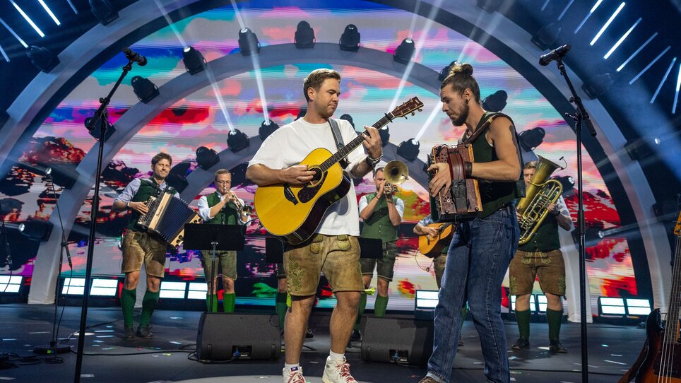 Ein Musiker spielt Gitarre, umgeben von einer Bläsergruppe und bunten Lichtern. | © Ivents Kulturagentur