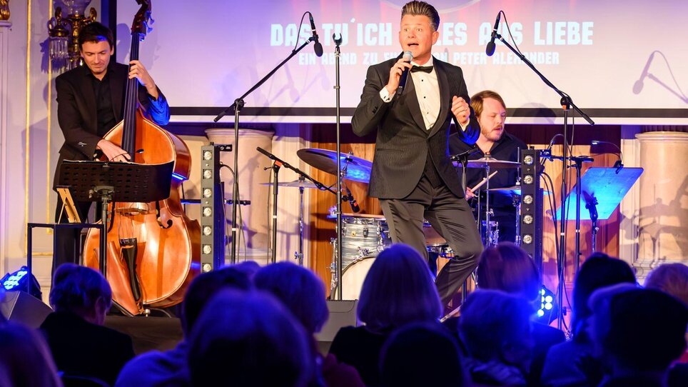 Ein Sänger performt, begleitet von Bass und Schlagzeug, während das Publikum zuschaut. | © Ivents Kulturagentur