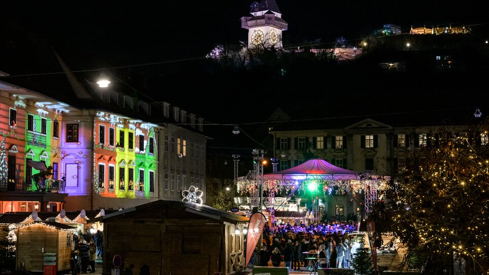 Bunte Lichter und Stände auf dem Weihnachtsmarkt in Graz, mit dem Grazer Uhrturm im Hintergrund. | © Ulrike Rauch