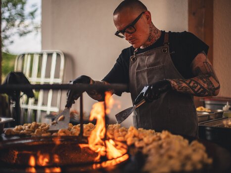 Ein Koch von The Underground Kitchen // Crew arbeitet konzentriert am Grill mit Flammen und zubereiteten Speisen. | © Alex Rauch