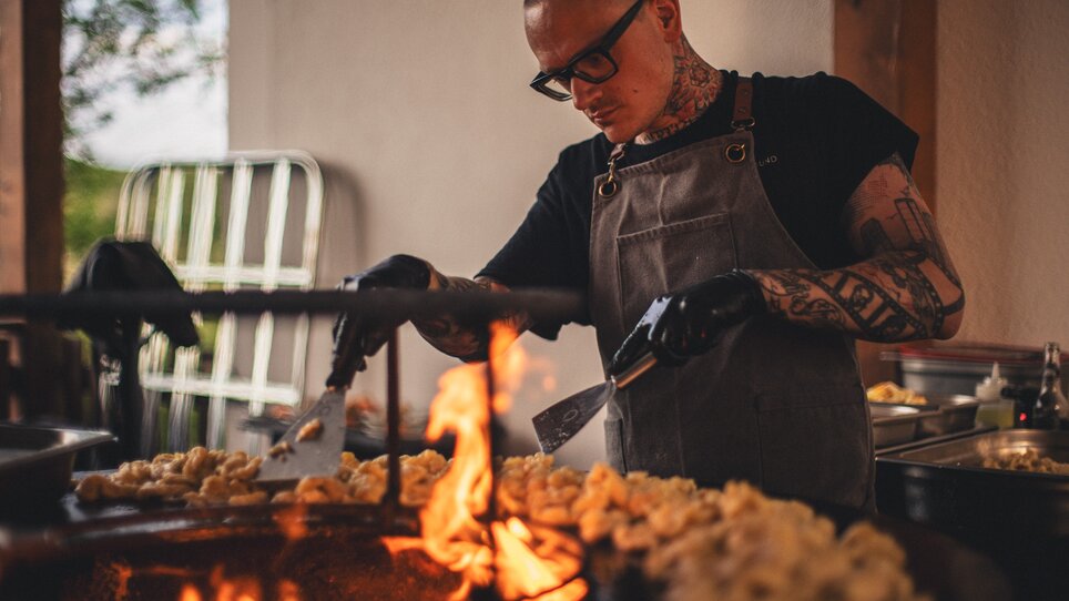 Ein Koch von The Underground Kitchen // Crew arbeitet konzentriert am Grill mit Flammen und zubereiteten Speisen. | © Alex Rauch