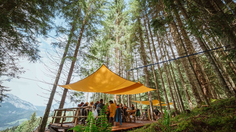 Eine Gruppe bespricht sich unter einem großen Sonnensegel in einem Wald. Der Veranstaltungsort hat Holzmöbel und ist gut beleuchtet. | © Alex Rauch