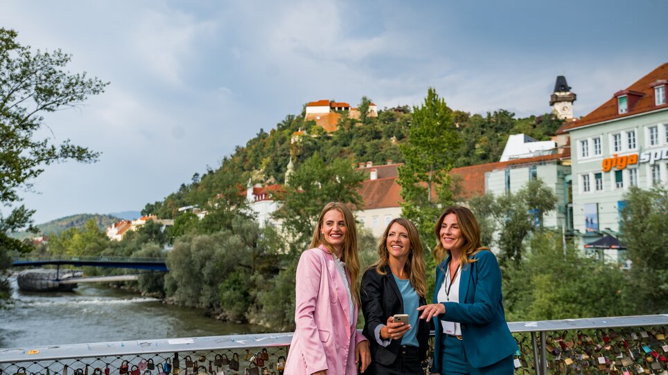 Drei Frauen posieren am Fluss Mur in Graz, mit Grazer Uhrturm im Hintergrund. | © Graz Tourismus - Mias Photoart