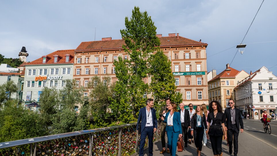 A group of businesspeople walk across a bridge in Graz, with historical buildings and trees in the background. | © Graz Tourismus - Mias Photoart