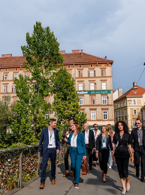 Eine Gruppe von Geschäftsleuten geht über eine Brücke in Graz, im Hintergrund historische Gebäude und Bäume. | © Graz Tourismus - Mias Photoart