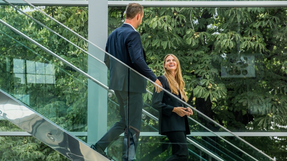 Zwei Personen in formeller Kleidung gehen eine Treppe in der Messe Graz hinauf, umgeben von moderner Architektur und Grün. | © Graz Tourismus - Mias Photoart