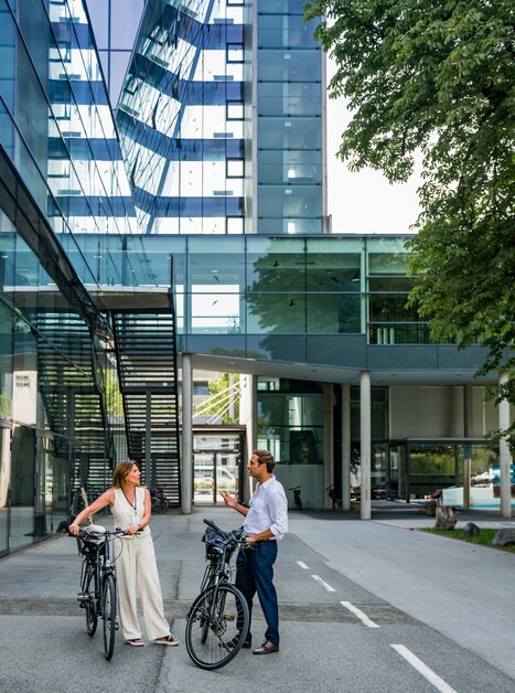 Two business-attired individuals converse while holding bicycles in front of modern architecture, Messe Graz. | © Graz Tourismus - Mias Photoart