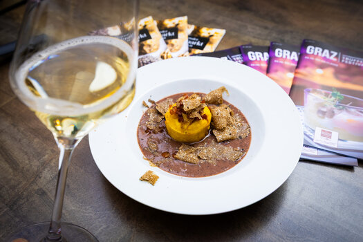 Trüffelfestival Graz - ein Trüffelgericht auf einem weißen Teller, ein Glas Weißwein und Prospekte vom Trüffelfestival auf einem braunen Tisch. | © Graz Tourismus - Werner Krug