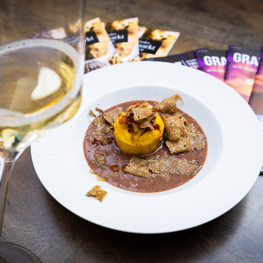 Trüffelfestival Graz - ein Trüffelgericht auf einem weißen Teller, ein Glas Weißwein und Prospekte vom Trüffelfestival auf einem braunen Tisch. | © Graz Tourismus - Werner Krug
