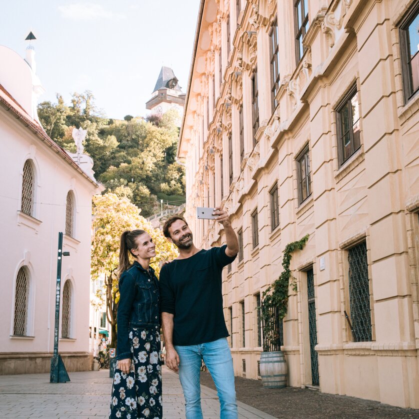 Una coppia sta facendo un selfie nella città di Graz, con la Torre dell'Orologio di Graz sullo sfondo. | © Graz Tourismus