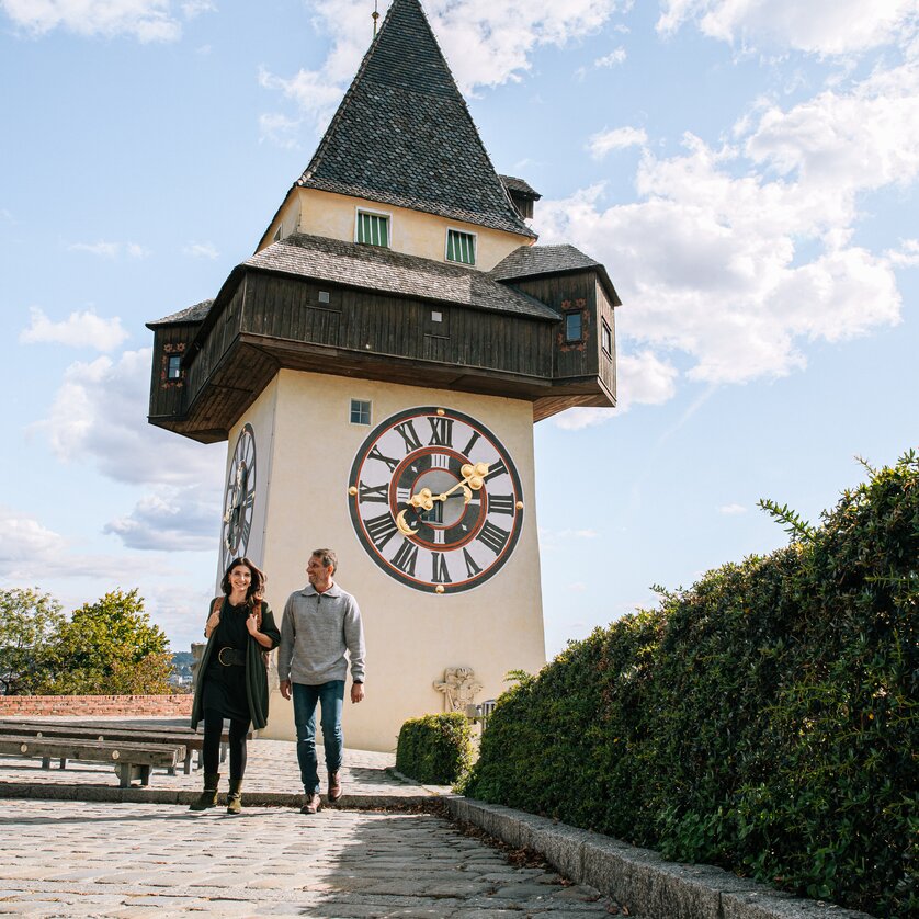 Coppia a passeggio davanti alla torre dell'orologio di Graz sotto un cielo azzurro. | © Graz Tourismus