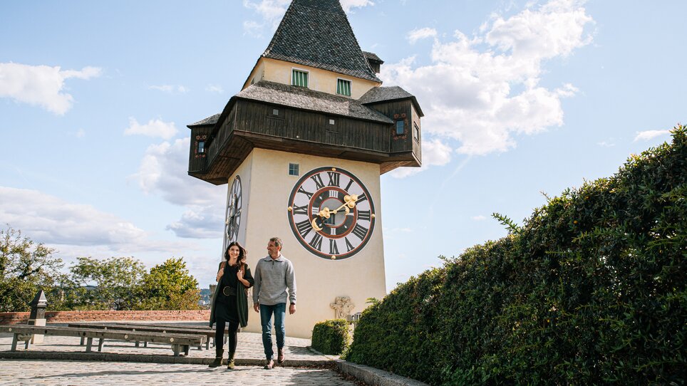 Couple walking in front of the Graz clock tower under a blue sky. | © Graz Tourismus