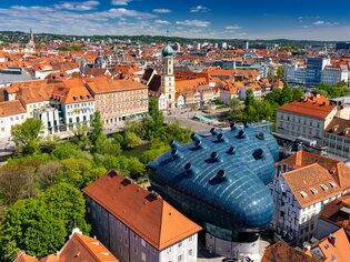 Blick auf Graz mit dem blauen Kunsthaus zwischen historischen Dächern. | © Graz Tourismus - Harry Schiffer