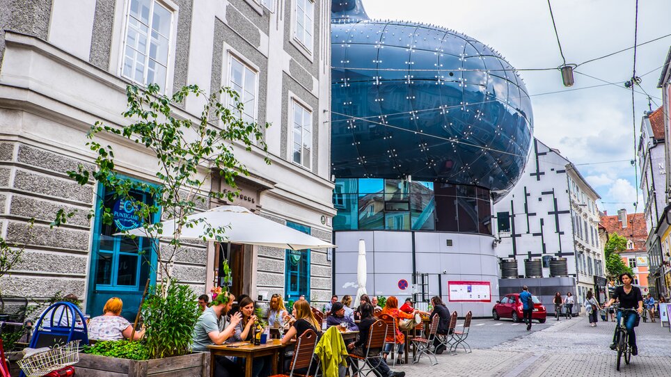 Blick auf das Kunsthaus Graz sowie ein Straßencafé mit Gästen. | © Becki Enright_Boarders of Adventure
