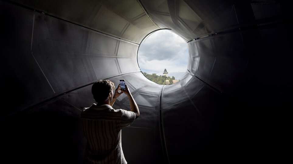 Besucher fotografiert den Grazer Uhrturm durch das Kunsthaus Graz. | © Graz Tourismus - Tom Lamm