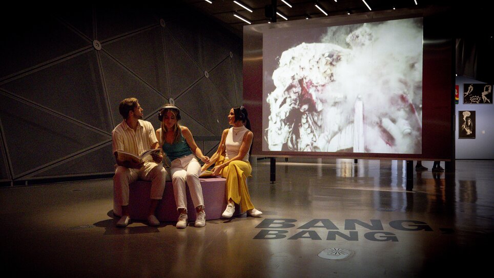 Drei Personen im Kunsthaus Graz, vor einem Video mit dem Text BANG. BANG. | © Graz Tourismus - Tom Lamm