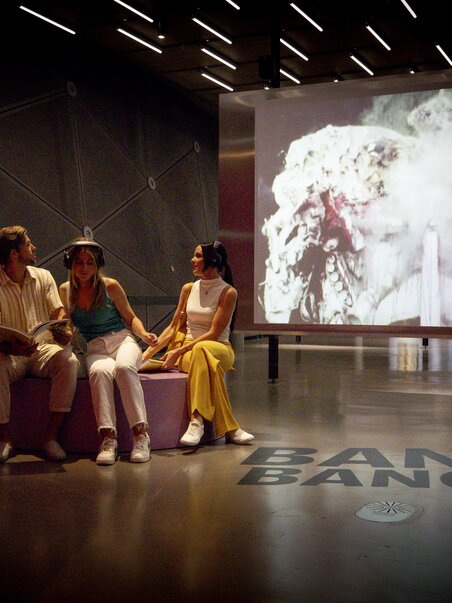 Three people in Kunsthaus Graz, in front of a video with the text BANG. BANG. | © Graz Tourismus - Tom Lamm