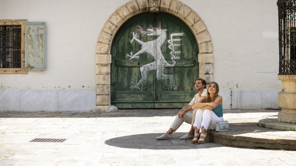 Couple sitting relaxed in Landhaus Courtyard in Graz in front of a colorful door. | © Graz Tourismus - Tom Lamm