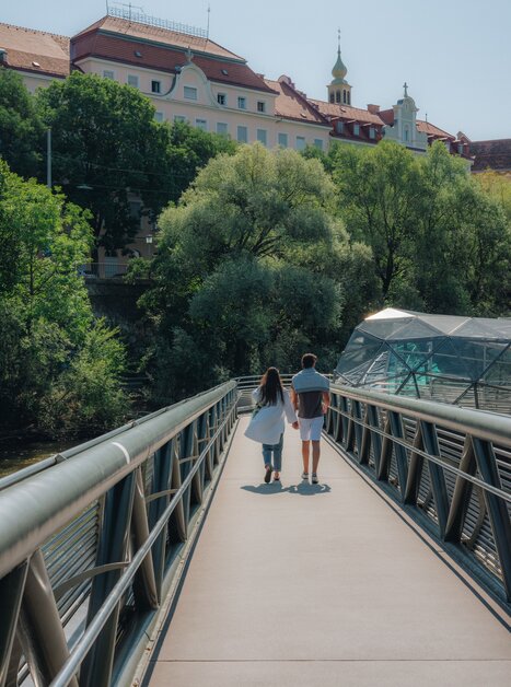 Ein Paar spaziert über die Murinsel in Graz, umgeben von Bäumen und Gebäuden.