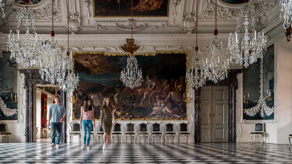 Three people walk through an opulent room with crystal chandeliers and wall paintings in Eggenberg Palace. | © Graz Tourismus - Mias Photoart