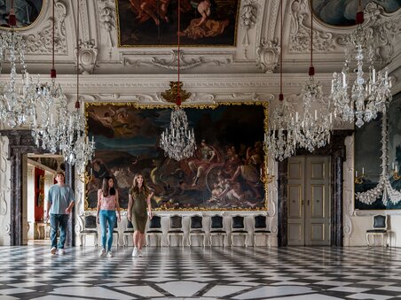 Three people walk through an opulent room with crystal chandeliers and wall paintings in Eggenberg Palace. | © Graz Tourismus - Mias Photoart