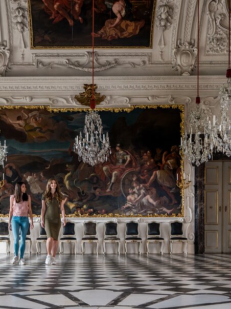 Three people walk through an opulent room with crystal chandeliers and wall paintings in Eggenberg Palace. | © Graz Tourismus - Mias Photoart