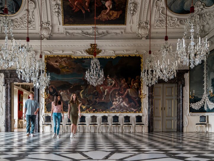 Three people walk through an opulent room with crystal chandeliers and wall paintings in Eggenberg Palace. | © Graz Tourismus - Mias Photoart
