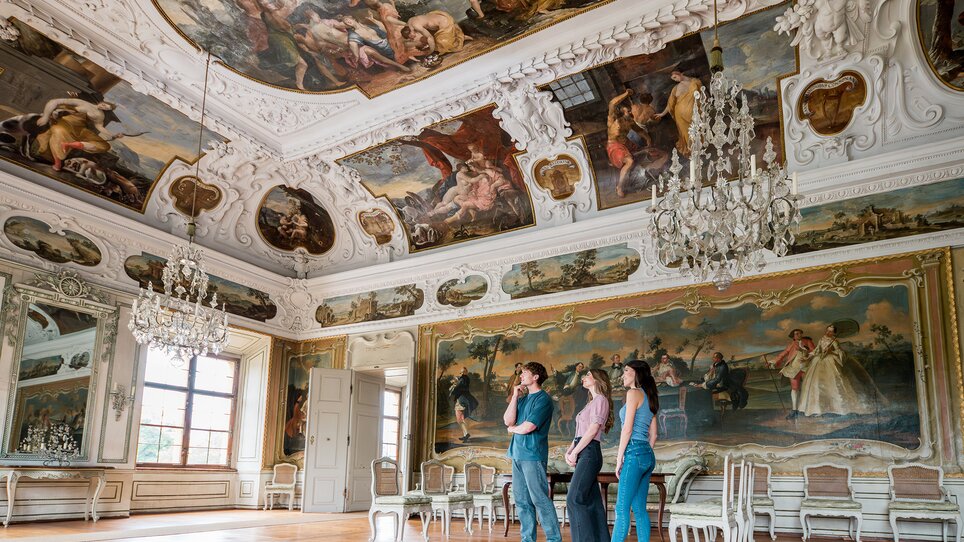 Drei Personen gehen durch einen opulenten Raum mit Kristallleuchtern und Wandmalerei im Schloss Eggenberg. | © Graz Tourismus - Mias Photoart