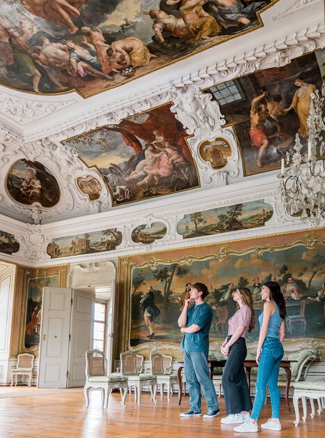 Three people walk through an opulent room with crystal chandeliers and wall paintings in Eggenberg Palace. | © Graz Tourismus - Mias Photoart