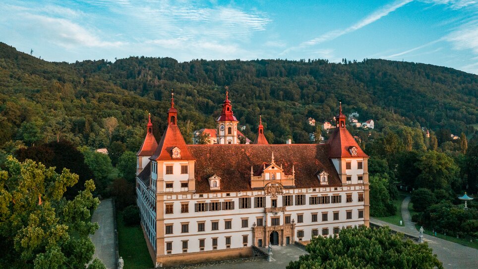 Ein Blick auf Schloss Eggenberg, umgeben von Bäumen und Bergen. | © Graz Tourismus - Mias Photoart