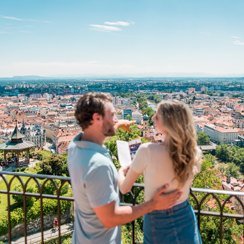 Ausblick vom Grazer Schlossberg | © Graz Tourismus - Mias Photoart Paar genießt den Ausblick auf Graz vom Schlossberg. Die beiden betrachten einen Prospekt von Graz. | © Graz Tourismus - Mias Photoart