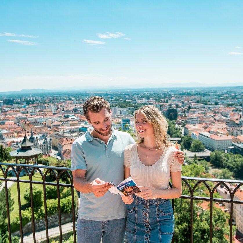 Paar genießt den Ausblick über Graz vom Schlossberg. Die beiden betrachten einen Prospekt von Graz. | © Graz Tourismus - Mias Photoart