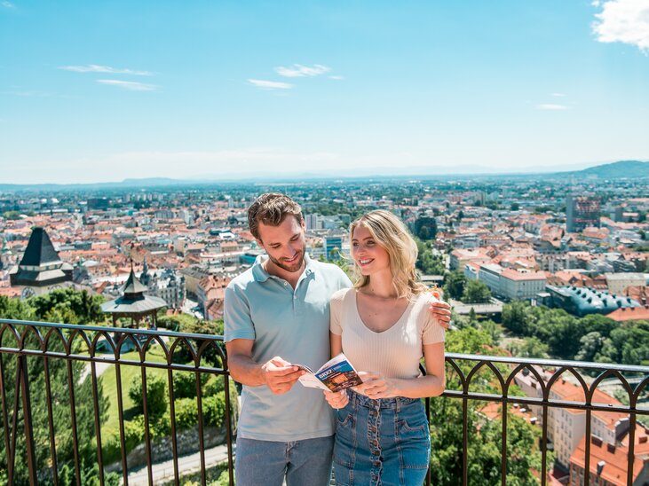 Couple enjoying the view over Graz from the Schlossberg. The two are looking at a brochure of Graz. | © Graz Tourismus - Mias Photoart